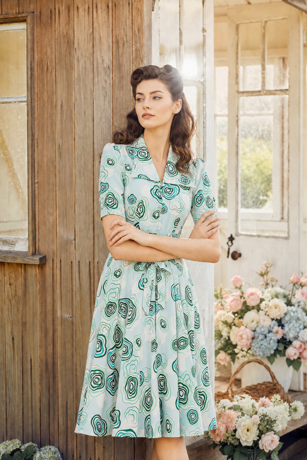 A woman stands in front of a rustic wooden door wearing a light blue knee-length vintage dress with a green and black spiral pattern. The setting includes a basket of pink and blue flowers on the right.