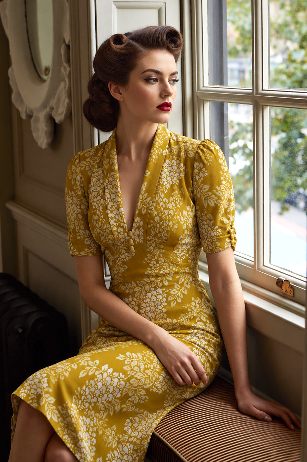 A woman with vintage-style curled hair sits by a window wearing a mustard yellow wrap dress with a white floral pattern. The photo is taken in an indoor setting with warm lighting.