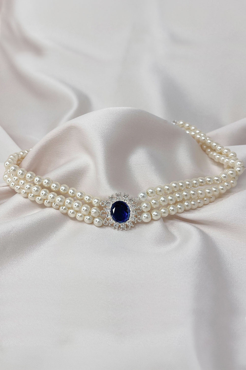 A double-row pearl necklace with a central oval dark blue gemstone surrounded by small clear stones, displayed on draped white satin fabric.